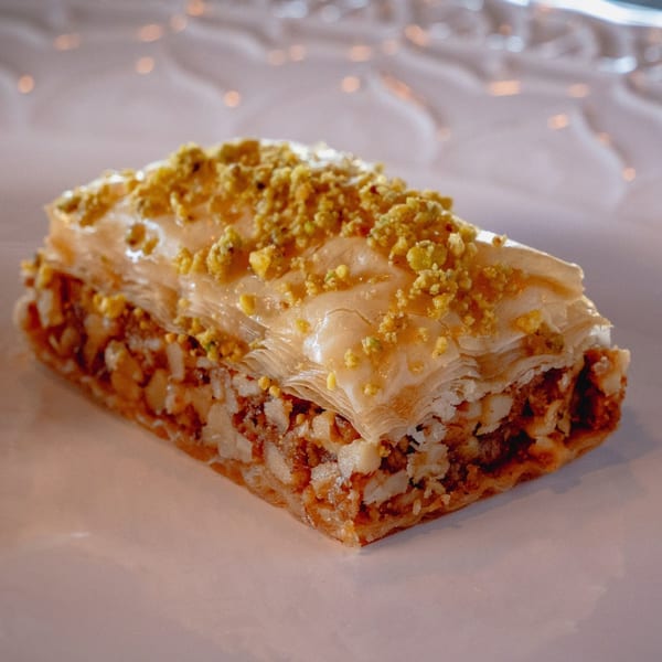 A close-up of a golden-brown baklava pastry topped with crushed pistachios on a white plate.
