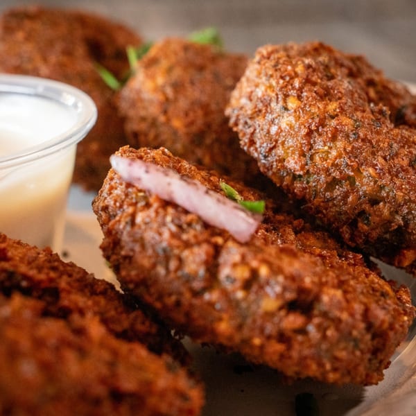 Close-up of crispy fried Mediterranean kibbeh, garnished with red onion and green herbs, with a side of sauce.