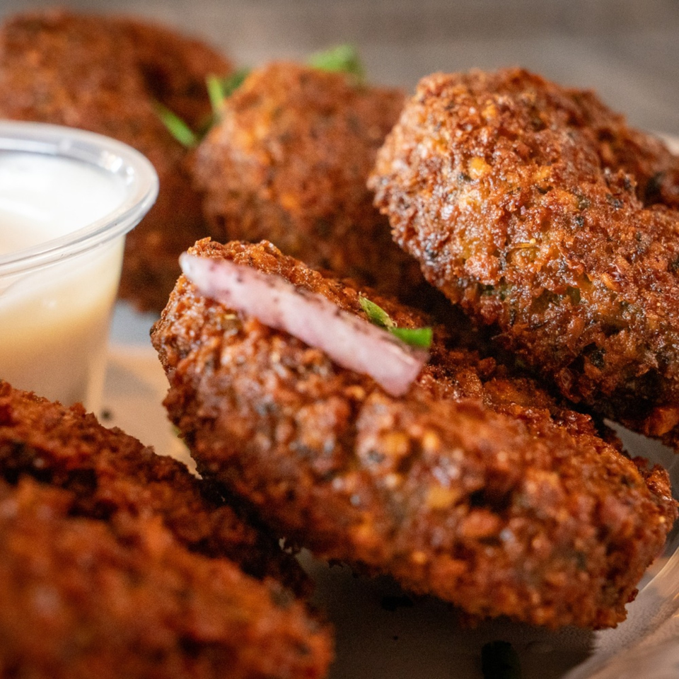 Close-up of crispy fried Mediterranean kibbeh, garnished with red onion and green herbs, with a side of sauce.